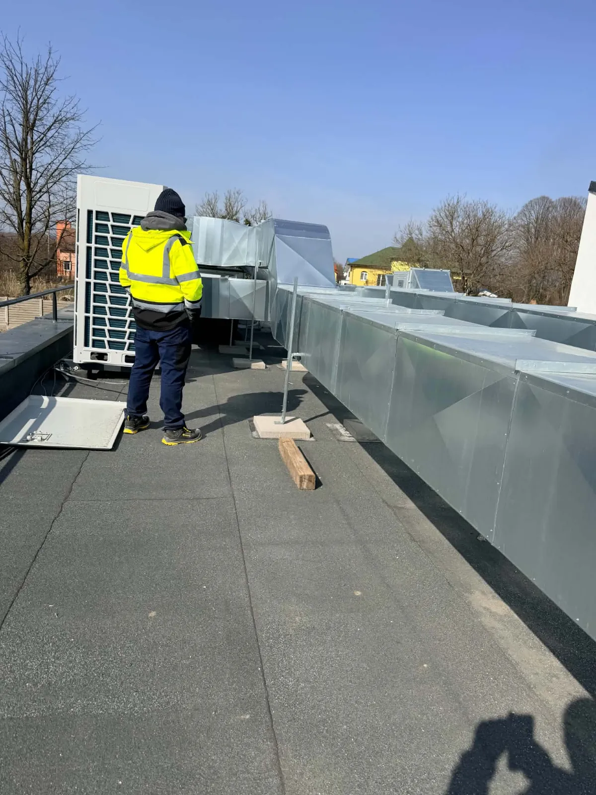 Tehnician în jachetă reflectorizantă lângă o unitate exterioară, pe un acoperiș plat, lângă un canal de ventilație zincat sprijinit pe suporți
