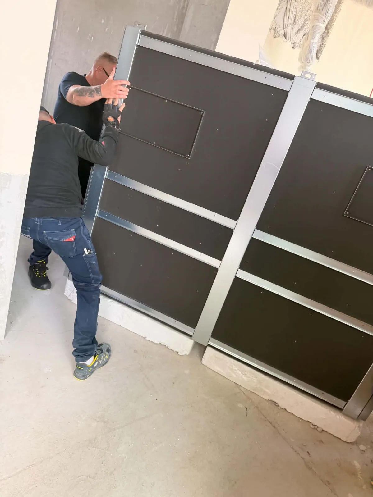 Doi oameni împing spre locul ei o unitate de tratare a aerului cu panouri negre și cadru zincat, așezată pe socluri de beton